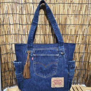 Op de afbeelding: Een donkerblauwe denim tote bag gemaakt van gerecyclede jeans, voorzien van een centrale achterzak, een Levi Strauss & Co. leren label, twee zijzakken en een bruine suède kwast met houten kralen bevestigd aan een van de twee schouderbanden.