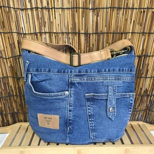 Op de afbeelding: Een blauwe denim schoudertas met een bruine leren riem en accenten. De tas heeft de zakken en details van een jeans, met een klein leren label met de tekst "ONE PASS". De tas staat op een houten oppervlak.