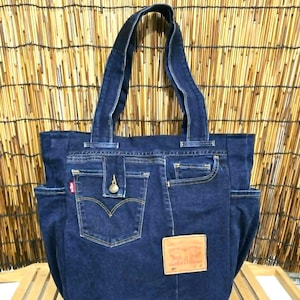 Puede incluir: Un bolso tote de mezclilla azul oscuro hecho de jeans reciclados. El bolso presenta los bolsillos originales, un cierre de botón y una etiqueta de cuero. El bolso tiene dos bolsillos laterales y asas resistentes.