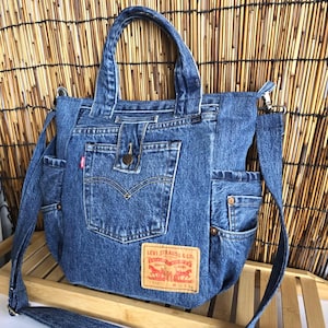 May include: Blue denim tote bag with a shoulder strap and two handles. The bag features multiple pockets and a Levi Strauss & Co. label. The bag is made from recycled denim and has a vintage look.