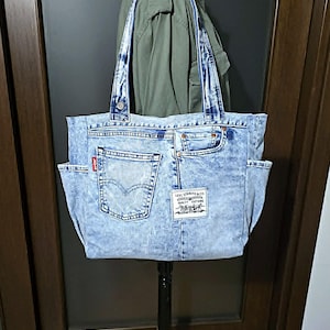 Puede incluir: Un bolso tote de mezclilla azul claro con lavado ácido, dos correas para el hombro, bolsillos laterales y un bolsillo trasero con etiqueta roja Levi's. El frente muestra un parche blanco de Levi Strauss & Co. con texto negro y dos caballos.