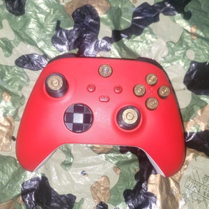 May include: A red video game controller with custom bullet casing buttons and thumbsticks. The controller has a checkered design on the center button and is set against a camouflage background.