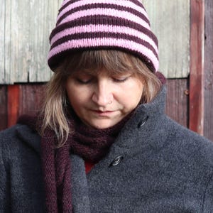 Puede incluir: Un gorro de punto con rayas horizontales rosas y marr&oacute;n oscuro. La persona lleva una bufanda morada oscura y un abrigo gris con botones negros grandes. El fondo es una puerta de madera.