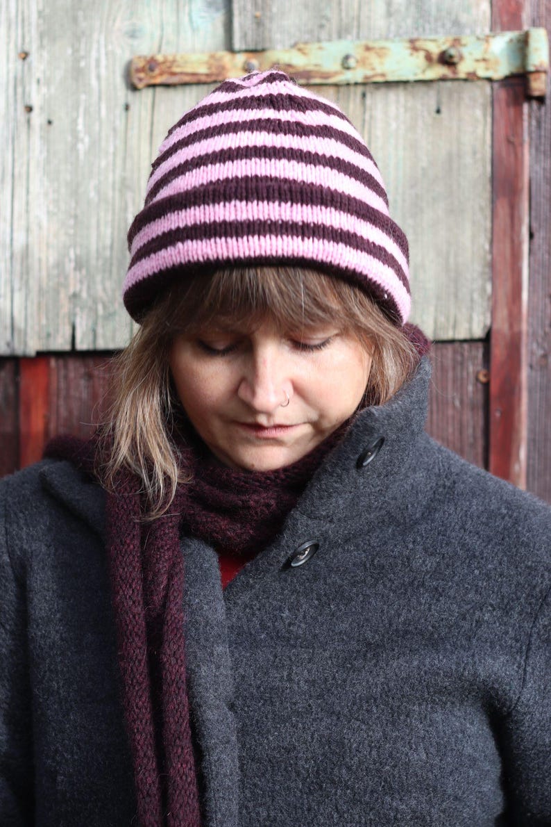 Puede incluir: Un gorro de punto con rayas horizontales rosas y marr&oacute;n oscuro. La persona lleva una bufanda morada oscura y un abrigo gris con botones negros grandes. El fondo es una puerta de madera.