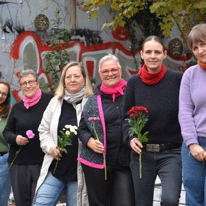 May include: A group of six people stand in front of a graffiti-covered wall, each holding a flower. They are wearing various outfits, including sweaters, jackets, and scarves in shades of pink, orange, yellow, and gray. The overall scene is outdoors.
