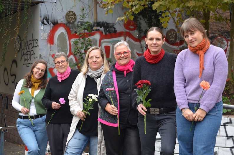 May include: A group of six people stand in front of a graffiti-covered wall, each holding a flower. They are wearing various outfits, including sweaters, jackets, and scarves in shades of pink, orange, yellow, and gray. The overall scene is outdoors.