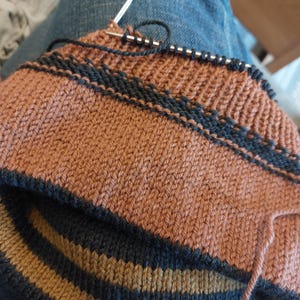 Puede incluir: Primer plano de un proyecto de tejido en curso. La pieza presenta un patr&oacute;n de rayas marr&oacute;n y azul marino. Una aguja de tejer sostiene varios puntos, con una madeja de hilo visible.