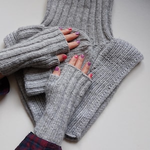 Puede incluir: Un cuello de punto gris claro y guantes sin dedos a juego. La prenda de punto acanalado se muestra con manos que usan los guantes. Los guantes tienen una abertura para el pulgar. El cuello está doblado y tiene un cuello alto. Las uñas están pintadas con esmalte de uñas rosa y morado.