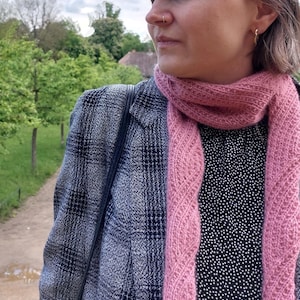 Könnte beinhalten: Ein rosa gestrickter Schal mit strukturiertem Design liegt über einem schwarz-weiß gepunkteten Oberteil. Der Schal wird mit einem dunkelgrauen Blazer mit Karomuster kombiniert. Der Hintergrund zeigt einen Weg und Bäume.