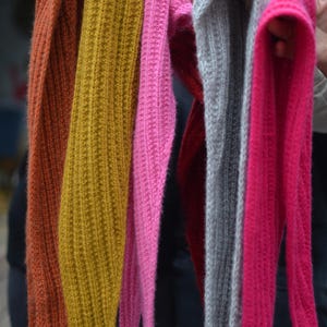 May include: A collection of colorful knitted scarves in shades of orange, mustard yellow, pink, red, gray, and fuchsia. The scarves are displayed vertically, showcasing their ribbed texture and varying lengths. The image highlights the variety of colors.