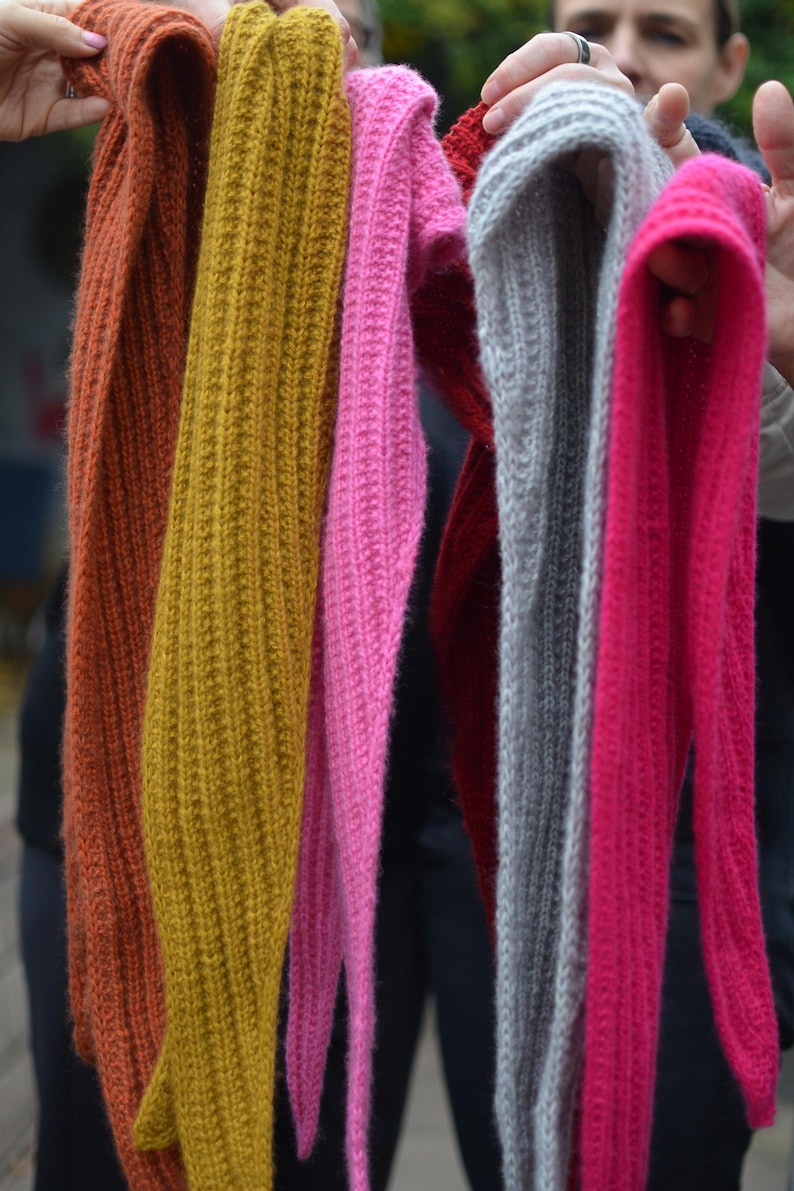 May include: A collection of colorful knitted scarves in shades of orange, mustard yellow, pink, red, gray, and fuchsia. The scarves are displayed vertically, showcasing their ribbed texture and varying lengths. The image highlights the variety of colors.
