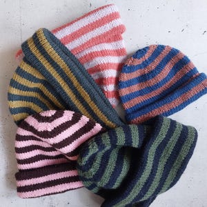 Könnte beinhalten: Eine Sammlung gestreifter Strickmützen und ein Schal in verschiedenen Farben. Die Mützen haben blau-korallenfarbene Streifen, rosa-braune Streifen und grün-marinefarbene Streifen. Der Schal hat rosa-weiße Streifen. Die Artikel sind auf einer hellgrauen Oberfläche angeordnet.