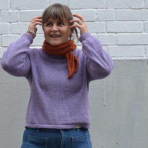 May include: A woman wearing a lavender sweater and blue jeans with large front pockets. She is also wearing an orange scarf. The background is a white brick wall and a gray wall.