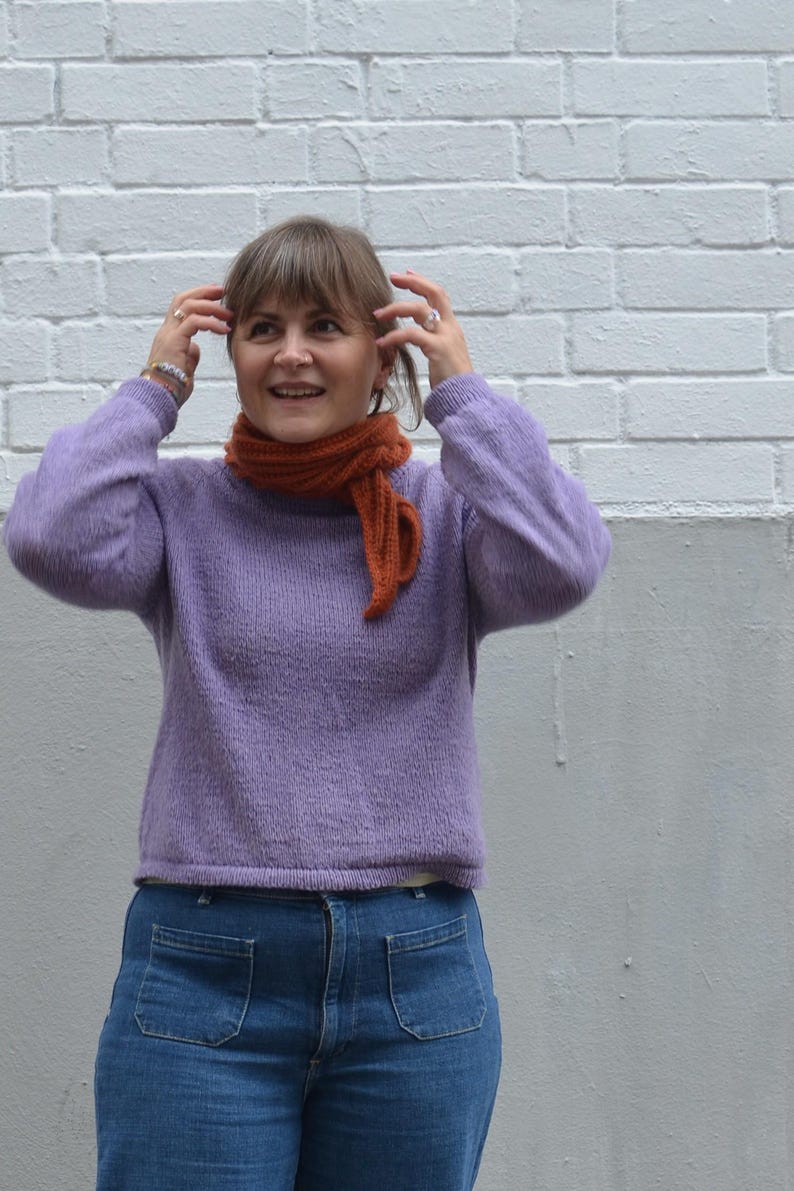 May include: A woman wearing a lavender sweater and blue jeans with large front pockets. She is also wearing an orange scarf. The background is a white brick wall and a gray wall.