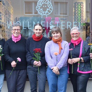 May include: Six people stand in front of a shop window, each holding a flower. They wear various sweaters, scarves, and jackets in colors like black, pink, and orange. The shop window displays yarn and the text "WOLLMEISTEREI".