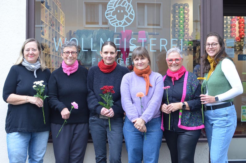 May include: Six people stand in front of a shop window, each holding a flower. They wear various sweaters, scarves, and jackets in colors like black, pink, and orange. The shop window displays yarn and the text "WOLLMEISTEREI".