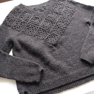 May include: A dark brown knitted jumper with a V-neck and long sleeves. The yoke has a geometric openwork pattern. The jumper is made from a soft, textured yarn, ideal for cooler weather.