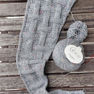 Puede incluir: Una bufanda gris tejida parcialmente terminada con un patrón de tejido de cesta, salpicada de motas coloridas. Una madeja de hilo gris y agujas de tejer están al lado de la bufanda. La etiqueta del hilo dice "Cardiff Cashmere". La bufanda está sobre una superficie de madera.