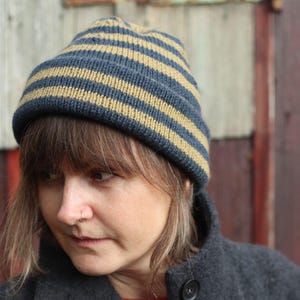 Puede incluir: Un gorro de punto a rayas azules y beige. La persona lleva un abrigo gris oscuro. El fondo es una pared de madera desgastada.