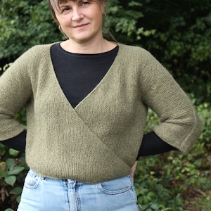 Könnte beinhalten: Ein olivgrüner Wickelpullover mit V-Ausschnitt. Der Pullover wird über einem schwarzen Oberteil getragen und mit hellblauen Jeans kombiniert. Die Ärmel sind dreiviertellang. Die Frau steht vor einem grünen, belaubten Hintergrund.