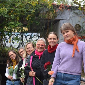 May include: A group of six people stand in a row, each holding a flower. They are wearing various outfits, including sweaters, jackets, and scarves. The background features greenery and graffiti.