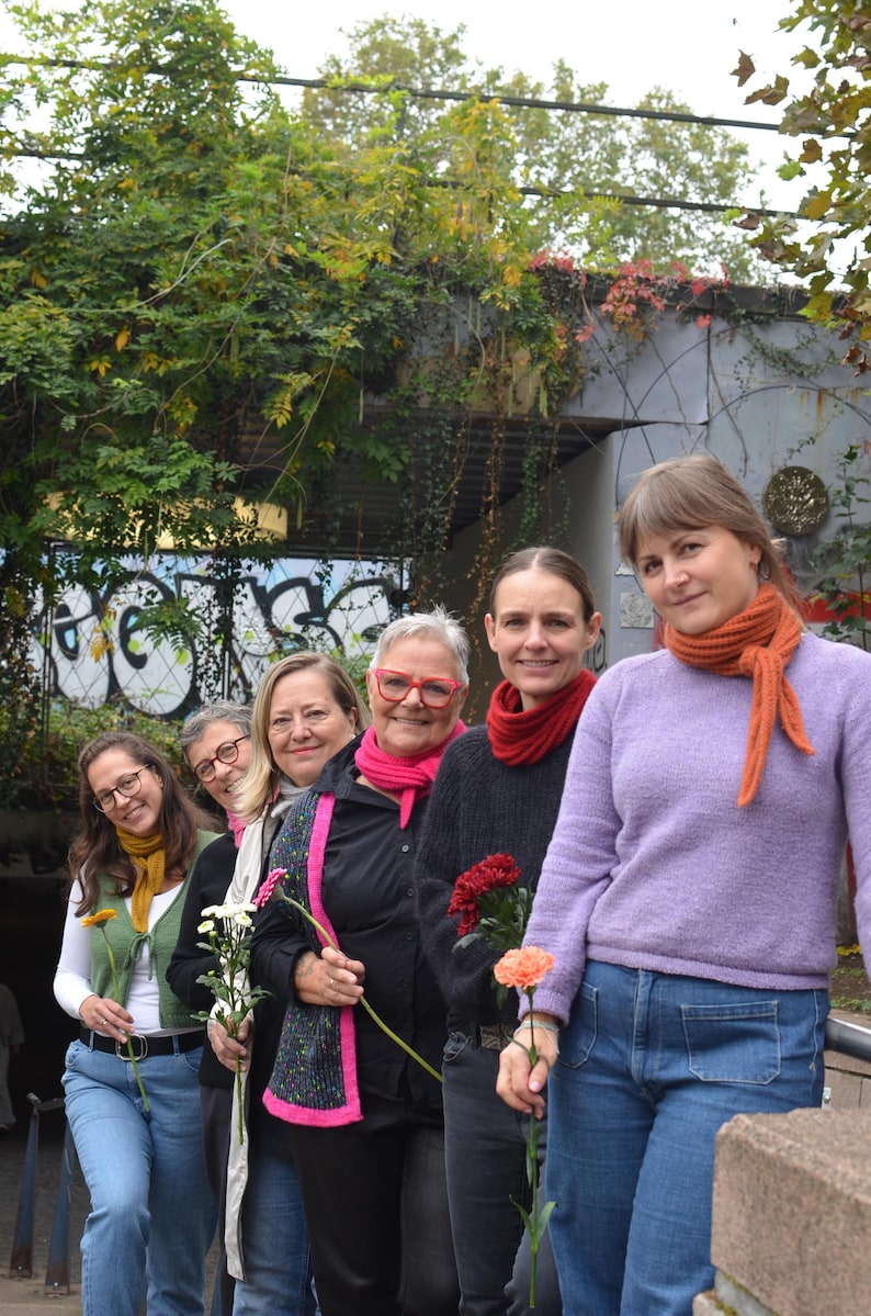 May include: A group of six people stand in a row, each holding a flower. They are wearing various outfits, including sweaters, jackets, and scarves. The background features greenery and graffiti.