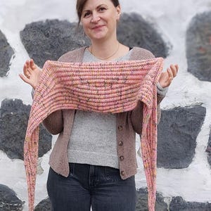 May include: A pink and white knitted scarf with a textured pattern. The scarf is draped around the neck of a person wearing a brown cardigan and jeans.