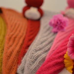 May include: Close-up of several knitted shawls in various colors, including mustard yellow, orange, red, gray, and pink. The shawls have a textured, ribbed design. Flowers are in the foreground.