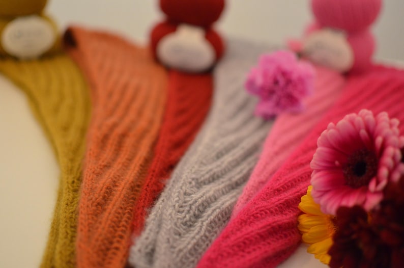 May include: Close-up of several knitted shawls in various colors, including mustard yellow, orange, red, gray, and pink. The shawls have a textured, ribbed design. Flowers are in the foreground.