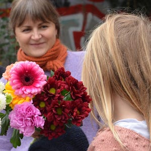 May include: A vibrant bouquet of flowers featuring a large pink gerbera daisy, deep red chrysanthemums, and other colorful blooms. The flowers are held by a person wearing a purple sweater, with another person in the background.