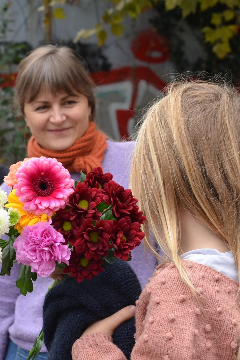 May include: A vibrant bouquet of flowers featuring a large pink gerbera daisy, deep red chrysanthemums, and other colorful blooms. The flowers are held by a person wearing a purple sweater, with another person in the background.