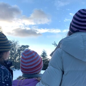 Puede incluir: Tres personas con gorros de punto a rayas y abrigos de invierno miran un paisaje nevado bajo un cielo nublado. Los gorros tienen rayas de varios colores, incluyendo azul, rosa y rojo. Los abrigos son blancos y morados.