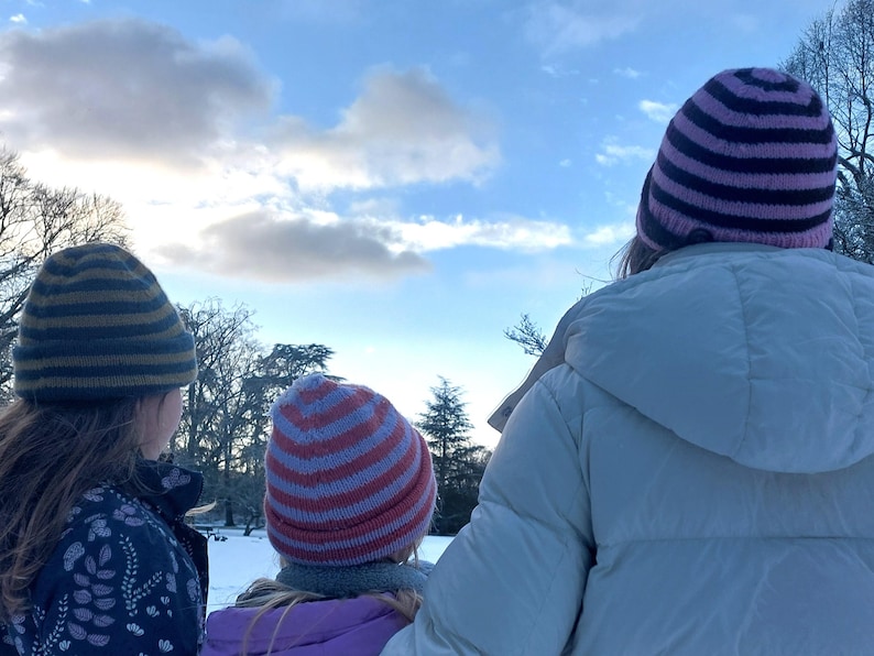 Puede incluir: Tres personas con gorros de punto a rayas y abrigos de invierno miran un paisaje nevado bajo un cielo nublado. Los gorros tienen rayas de varios colores, incluyendo azul, rosa y rojo. Los abrigos son blancos y morados.