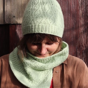 Könnte beinhalten: Ein hellgrünes Strickmützen- und Schal-Set. Die Mütze hat ein strukturiertes Muster, und der Schal ist dreieckig. Das Set ist aus weichem Garn gefertigt, ideal für kaltes Wetter. Der Hintergrund ist eine Holzwand.