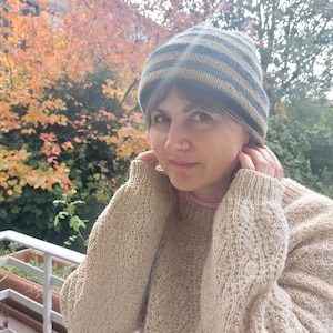 Puede incluir: Un su&eacute;ter de punto beige con un patr&oacute;n texturizado y un gorro de punto a rayas azul y beige. El gorro tiene rayas horizontales. La persona lleva una camiseta interior rosa.