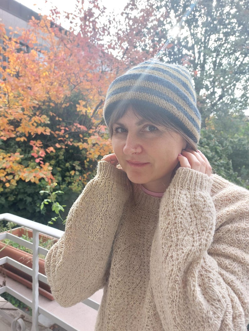 Puede incluir: Un su&eacute;ter de punto beige con un patr&oacute;n texturizado y un gorro de punto a rayas azul y beige. El gorro tiene rayas horizontales. La persona lleva una camiseta interior rosa.