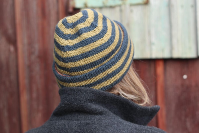 Puede incluir: Un gorro de punto con rayas alternas en azul marino y amarillo dorado. El gorro lo lleva una persona, de la que solo se ve la parte posterior de la cabeza y los hombros. La persona lleva un abrigo gris oscuro.