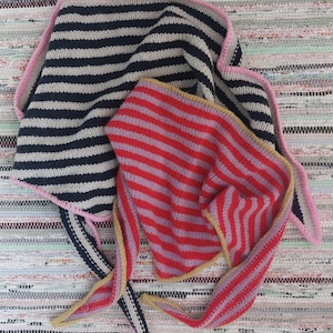 Può includere: Due scialli lavorati a maglia con righe orizzontali blu navy e crema, e rosso e lavanda. Lo scialle blu navy ha un bordo rosa. Lo scialle rosso ha un bordo giallo. Gli scialli sono su uno sfondo a righe multicolore.