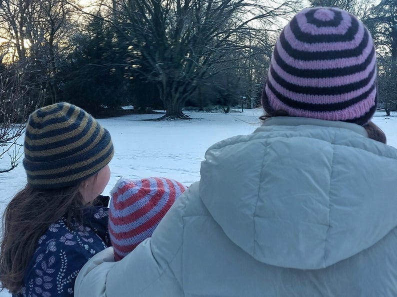 Puede incluir: Escena invernal con personas que llevan gorros de punto a rayas. Los gorros presentan combinaciones de colores como azul y dorado, rojo y blanco, y morado y negro. Una persona lleva una chaqueta con capucha blanca.