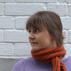 May include: A woman wearing a knitted orange scarf and a lavender sweater. The scarf is wrapped around the neck and tied in a knot. The background is a white brick wall.