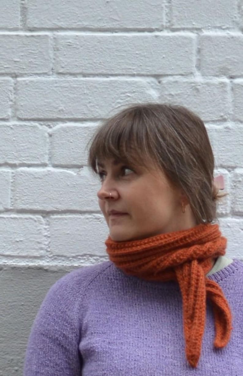 May include: A woman wearing a knitted orange scarf and a lavender sweater. The scarf is wrapped around the neck and tied in a knot. The background is a white brick wall.
