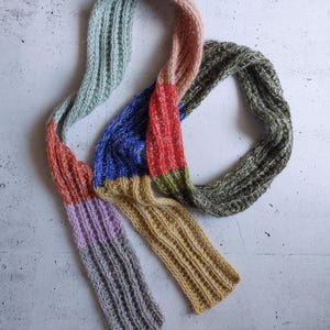 May include: A colorful, hand-knitted scarf with a ribbed texture. The scarf features sections of various colors, including blue, red, green, yellow, and gray. The scarf is made of soft yarn and is designed for warmth and style.