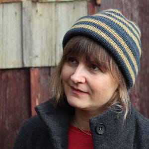 Puede incluir: Un gorro de punto a rayas azul marino y beige. La persona lleva un abrigo gris oscuro y una camiseta roja. El fondo es una pared de madera desgastada.