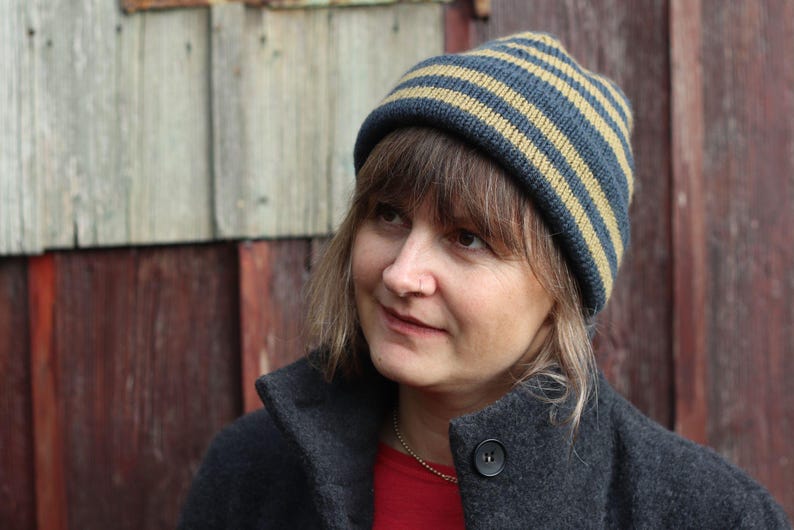 Puede incluir: Un gorro de punto a rayas azul marino y beige. La persona lleva un abrigo gris oscuro y una camiseta roja. El fondo es una pared de madera desgastada.