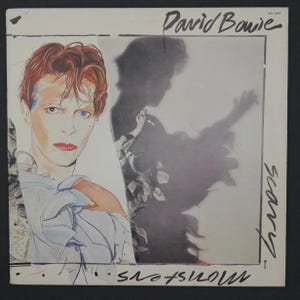 May include: Album cover for David Bowie's "Scary Monsters" album. The artwork shows a portrait of Bowie with red hair and a blue eye, next to a shadowy figure. The album title and artist's name are handwritten in black ink.