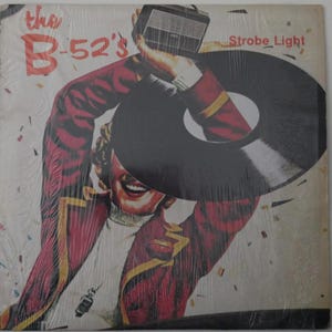 May include: Album cover for The B-52's "Strobe Light" record. The artwork features a person in a red jacket holding a vintage radio and a vinyl record. The band's name is in red, and the album title is in white.