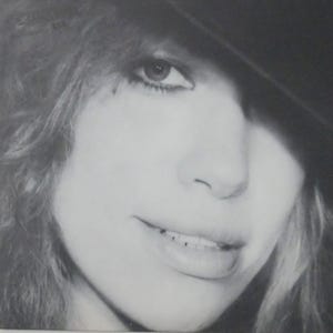 Puede incluir: Retrato en blanco y negro de una persona con cabello largo y ondulado y un sombrero. La persona mira hacia la cámara con una ligera sonrisa. La imagen es un primer plano, centrado en la cara y la parte superior del cuerpo.
