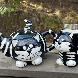 May include: A whimsical black and white cat-themed teapot and matching mug set. The teapot features a striped design with a cat face and paw feet. The mug mirrors the design, creating a charming tea set.