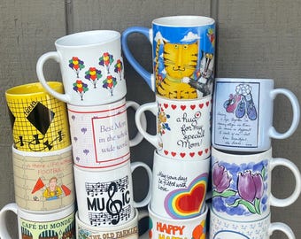 May include: A collection of various ceramic mugs with colorful designs. Some mugs feature text like "Happy Birthday!" and "Best Mom." Other designs include balloons, musical notes, a cat, and floral patterns. The mugs are stacked on top of each other.