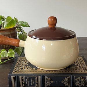 May include: Cream-colored, lidded soup pot with a brown rim and lid, and a wooden handle. The pot sits on a decorative book, with a potted plant in the background. The pot is a classic kitchenware item.